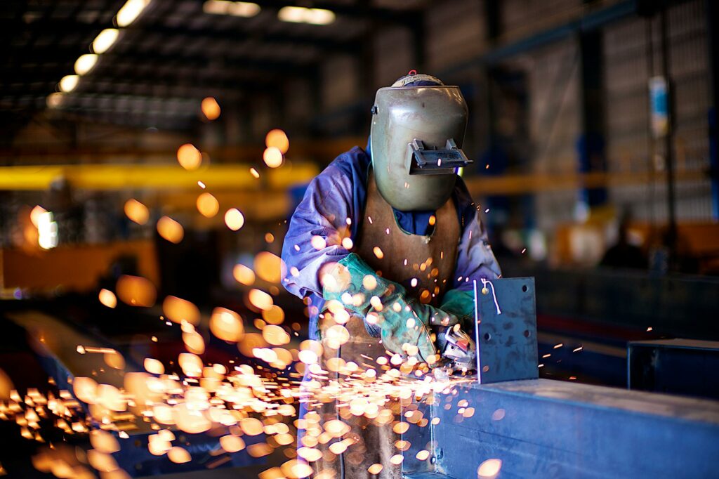 Person (m/w/d) mit Schutzhelm und Schweißgerät, Funkenflug bei der Arbeit an einem Metallbauteil.